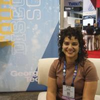 Lorna Rivera sits on a white couch in front of a pale blue banner at SC'19 in the Georgia Tech booth.